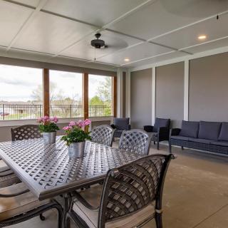 A covered patio with a dining table, chairs, and potted flowers, featuring large windows and a ceiling fan.