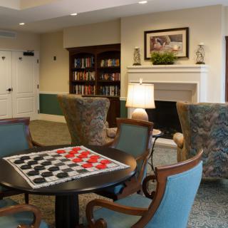 A cozy living room featuring a chessboard on a table surrounded by comfortable chairs, with a bookshelf and fireplace in the background.