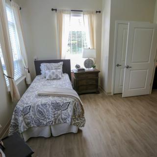 A cozy bedroom featuring a single bed with patterned bedding, a nightstand with a lamp, and windows with sheer curtains.