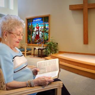 An elderly woman reads a Bible in a church with stained glass and a cross in the background.