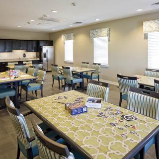 A spacious community dining and activity area with tables, chairs, and a kitchen in the background.