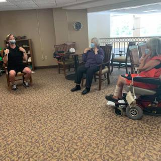 A group of seniors sitting in a common area, some wearing masks and one in a wheelchair.