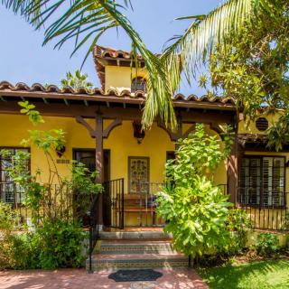 Charming Mediterranean-style house with a yellow exterior, surrounded by lush greenery and palm trees.