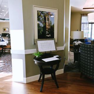 A lobby area with a small table featuring a plant and a framed sign, alongside wall art and comfortable seating.