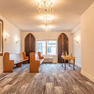 A small chapel room featuring wooden benches, chandeliers, and arched windows.