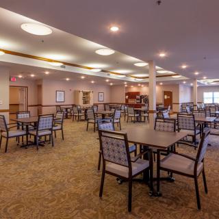 A spacious dining hall with multiple tables and chairs, featuring a carpeted floor and well-lit interior.