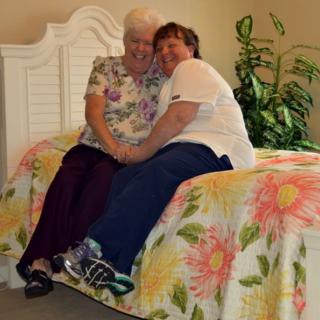Two people sharing a warm embrace while sitting on a bed inside a cozy room.