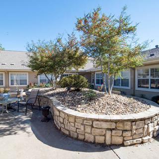 A serene courtyard featuring outdoor seating, surrounded by trees and a stone wall under a clear blue sky.