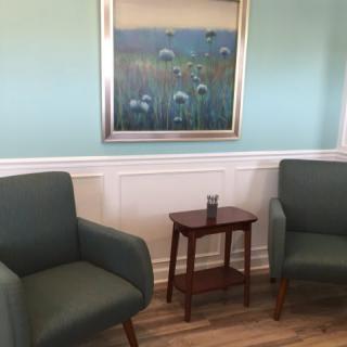 A cozy waiting room with two green chairs and a wooden table beneath a floral painting.
