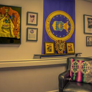 An office space featuring a flag, framed memorabilia, and a couch with colorful cushions.