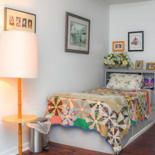 A cozy bedroom with a single bed adorned with a colorful quilt, surrounded by artwork and soft lighting.