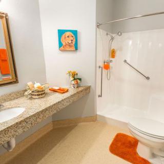 A well-decorated and accessible bathroom with a shower, toilet, and sink, featuring orange accents and artwork on the wall.