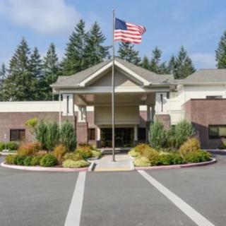A building entrance with an American flag on a pole, surrounded by landscaped greenery and trees under a clear blue sky.