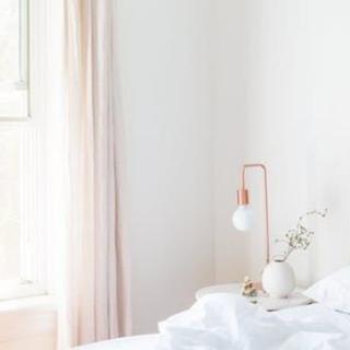 A minimalist and bright bedroom interior with a white color scheme, featuring a modern lamp and decorative vase on the bedside table.