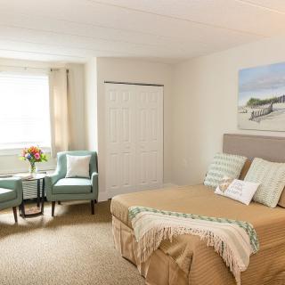 A cozy bedroom interior featuring a bed, two chairs by the window, and soft decor elements.