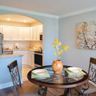 A modern dining area with a round table set for two, adjacent to an open kitchen and decorated with a potted plant and artwork on the wall.