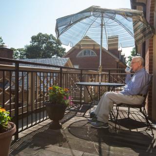 A man enjoys a sunny day on a balcony, sitting under an umbrella with potted plants nearby.