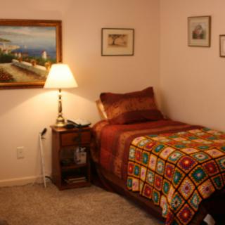 A cozy bedroom with a single bed adorned with a colorful quilt, a nightstand with a lamp, and various paintings on the walls.