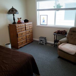 A cozy bedroom with a dresser, armchair, and a bed, illuminated by natural light from the window.