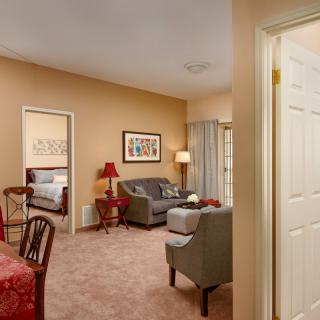 A cozy living room with a sofa, armchair, and table, leading into a bedroom.