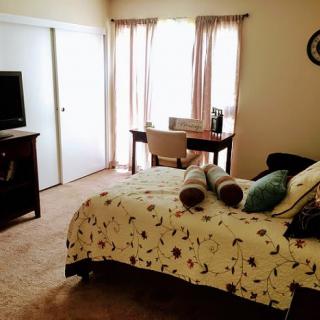 A cozy bedroom featuring a neatly made bed, a television, and a desk near the window with curtains.