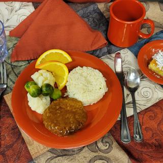 A meal served on a dining table, featuring rice, a meat patty, Brussels sprouts, orange slices, with a cup and dessert cake on an orange-themed setting.