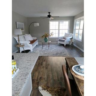 A modern living room with a neutral color palette featuring a couch, armchair, coffee table, and hardwood flooring, illuminated by natural light from large windows.