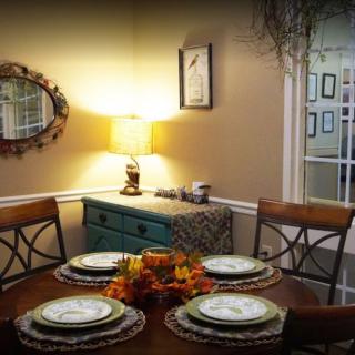 A cozy dining room with a round table set for four, featuring a lit lamp and decorative mirror.