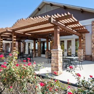 A sunny patio with a wooden pergola and outdoor seating, surrounded by blooming flowers.