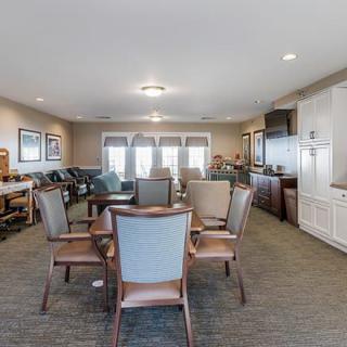 A spacious living room with chairs and tables arranged around a central area, featuring large windows and warm decor.