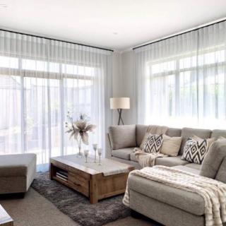 A cozy living room with a sectional sofa, wooden coffee table, and large windows draped with sheer curtains.