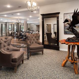A stylish lobby area featuring tufted armchairs, a unique table sculpture, and a large mirror, with a view into an elegant dining area.