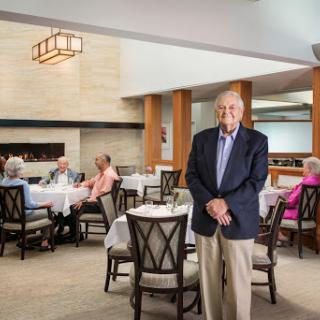 An elderly gentleman stands in a well-lit restaurant, surrounded by other diners enjoying their meals and conversations.