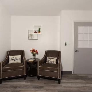 A minimalist living room corner featuring two brown armchairs with decorative pillows, a small round table with a vase of flowers, and a wall-mounted shelf.
