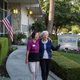 A caregiver and an elderly woman walk arm in arm outside a senior living community.