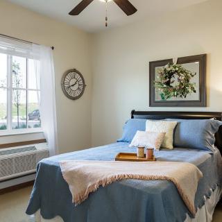A cozy bedroom with a neatly made bed, decorative pillows, and a wall clock, complemented by a large window providing natural light.