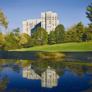 A tall building surrounded by trees reflected in a calm lake under a clear blue sky.