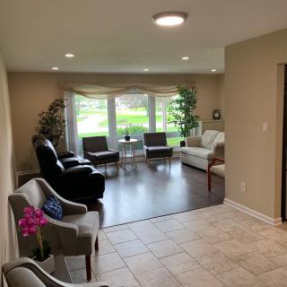 A cozy living room with ample natural light, featuring comfortable seating and a view of greenery through large windows.