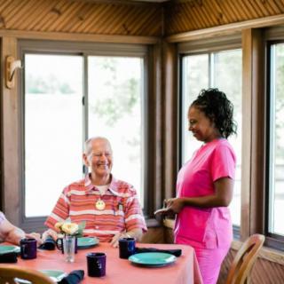 A caregiver in pink scrubs interacts with elderly residents during a meal in a nursing home setting.