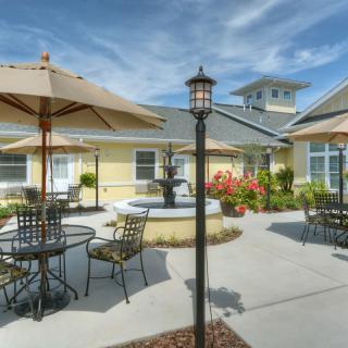 A sunny outdoor patio area featuring tables and chairs with umbrellas, a central fountain, and well-maintained landscaping.