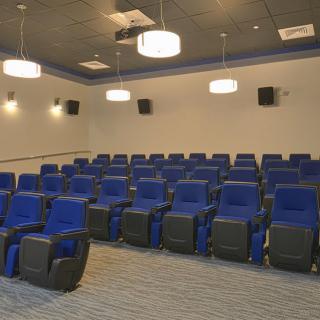 An empty theater room with rows of blue seats and overhead lighting.