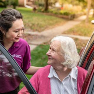 A caregiver assisting an elderly person getting out of a car in a garden setting.
