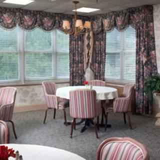 A cozy dining room with round tables and striped chairs, adorned with floral curtains and a hanging chandelier.