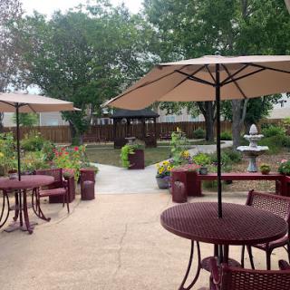 A serene outdoor garden space with tables under umbrellas, a pathway leading to the fountain, and lush greenery.