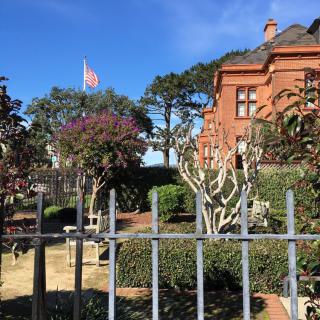 A brick building with a garden, trees, and a flag on display behind a metal fence on a sunny day.