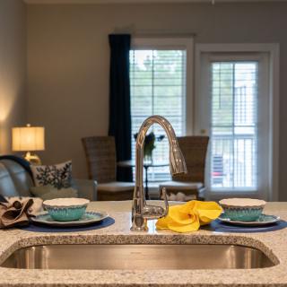 A modern kitchen with a view of the dining area through a window, featuring a sleek faucet and neatly arranged tableware on the countertop.