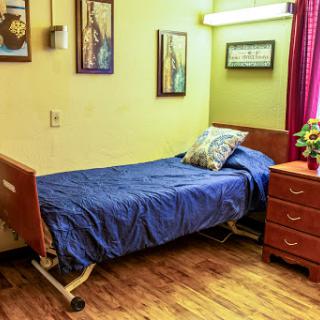 A cozy hospital or nursing home room featuring a bed with blue bedding, a wooden dresser, and decorative artwork on the walls.