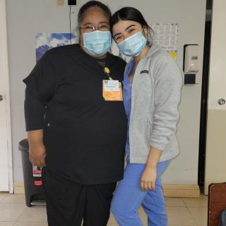 Two healthcare workers wearing masks and uniforms pose for a photo in a hospital setting.