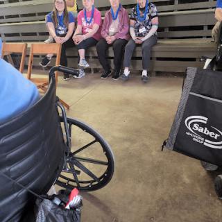 A group of individuals wearing medals seated together in an indoor setting, with a wheelchair in the foreground.