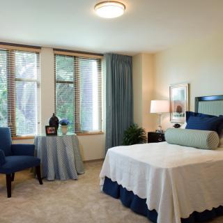 A cozy bedroom with a neatly made bed, a blue chair, and large windows covered with curtains.